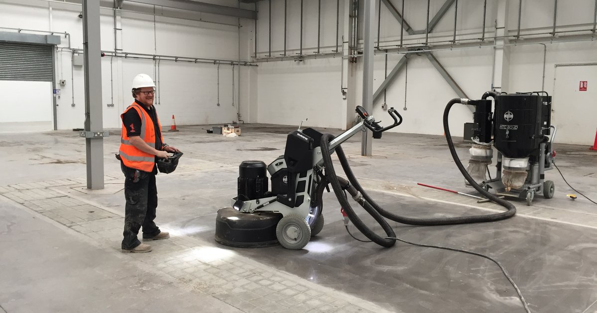 Contractor operating a walk-behind diamond floor grinder connected to a HEPA dust extractor on a bare concrete slab